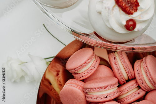  An elegant pairing of pastel pink macarons filled with vanilla cream alongside white meringue desserts topped with whipped cream, raspberries, and red currants. Perfect for a wedding or upscale event