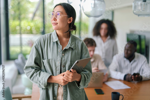 A joyful Asian young adult businesswoman holds a digital tablet, looking content in a buzzing modern office with diverse colleagues in the background.