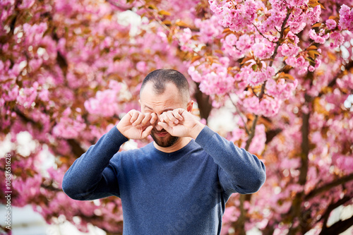 Man allergic suffering from seasonal allergy at spring in blossoming garden at springtime. Bearded man feeling itchy eyes in front of blooming tree. Spring allergy concept.