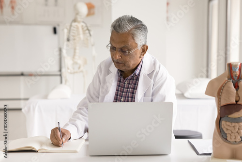 Elderly Indian male doctor dressed in white coat sit at workplace desk with laptop writing information, makes note in register, filling patient anamnesis in medical journal, working in clinic office