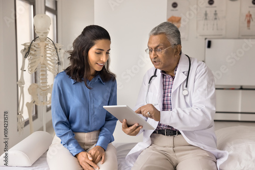 Older Indian GP explains treatment plan, showing check up health test results on digital tablet to young female patient at her visit in clinic, smile, receive professional medical services at hospital