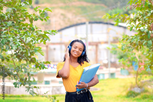 Universitária negra feliz a caminho da faculdade enquanto escuta música nos fones de ouvido sem fio. Ela está numa praça com árvore, sorrindo e com olhos fechados. 