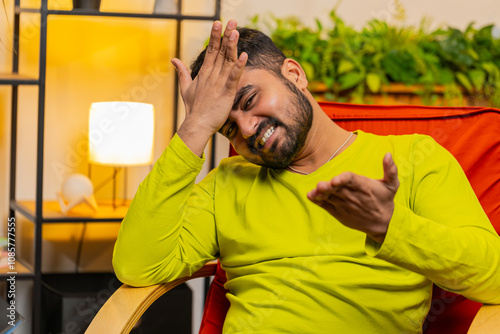 Face palm. Shame on you. Upset Indian man making disrespect gesture, feeling bored, disappointed in fail results, bad news. Unhappy confused Arabian Hindu guy at home apartment room sitting on chair