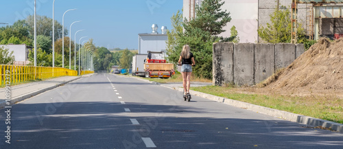 Droga w byłej dzielnicy przemysłowej pod błękitnym niebem. Piękny, słoneczny poranek późnego lata w mieście - asfaltową drogą przez „poprzemysłową” dzielnicę miasta jedzie dziewczyna na e hulajnodze .