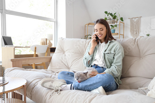 Young woman with cute British Shorthair cat talking by mobile phone on sofa at home