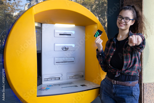 Young white girl withdraws money from an ATM while posing with her credit card and points at the camera with her finger