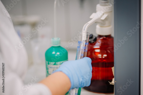 A gloved hand filling a graduated cylinder with liquid from a dispenser in a laboratory, with bottles visible in the background.