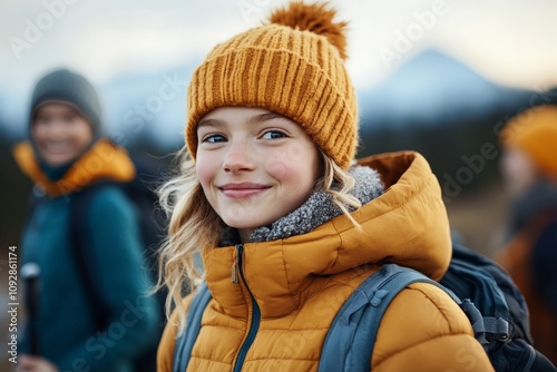 A young girl smiling warmly in a knitted hat and jacket, standing amidst snowy mountains, embracing the joys of a winter hiking adventure.