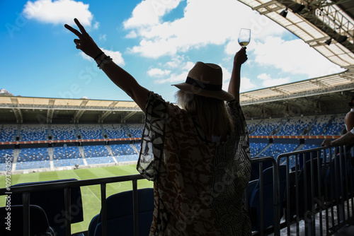 A woman with a hat raises her hands to the sides against the background of a football stadium and a cloudy blue sky