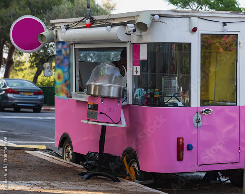 A bright pink ice cream cart, parked on a quiet street under the shade of green trees with playful designs. The scene captures a carefree and inviting summer afternoon, perfect for a cool snack