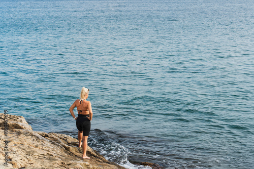 A slender blonde girl stands on the rocky shore of the Mediterranean Sea on a summer day and gets ready to swim.