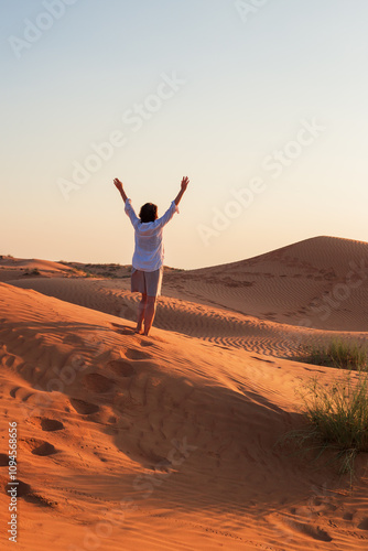 Happy woman is standing with open arms at sand dune and enjoying sunset.