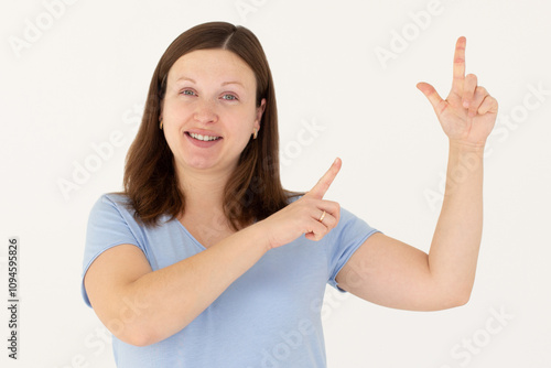 Hey look. Smiling pretty female model pointing fingers right at empty space and inviting to check it out, showing advertisement on white background.