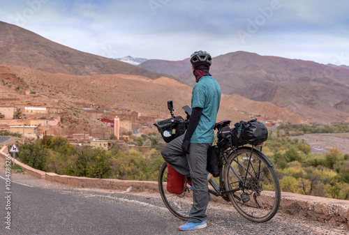 Bike packer man in Atlas Mountains in Morocco, admires village with mosque, colorful mountains and snow caps. Nomad traveling on his bike.