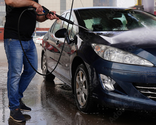 A muscular man dressed in a black T-shirt and blue jeans washes his blue-colored car at a touchless car wash