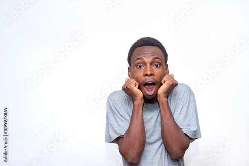 A young man in a grey t-shirt shows a surprised expression with hands on cheeks. His shocked face conveys astonishment and surprise against a plain white background, highlighting his emotions.