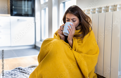 Woman trying to warm with hot cup of tea, covered with blanket next to radiator