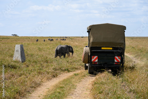 Safari stopped vehicle on dirt road to let an elephant pass in front, Tanzania Kenya border marker stone on savanna, African safari vacation in Kenya, Maasai Mara National Reserve