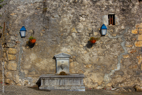 Old stone fountain dated 1811 on a rustic wall in an Italian village, decorated with lanterns and flower pots, evoking timeless charm.