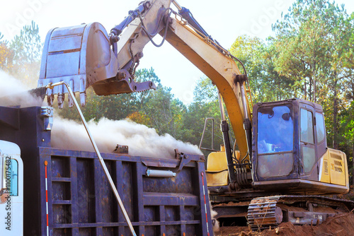 Under land removal, an excavator transfers large quantities of dirt into truck, creating dust clouds in construction area