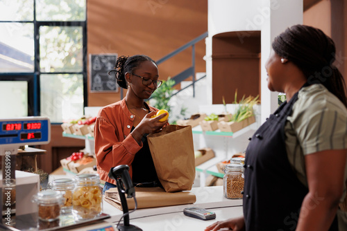 African American woman vendor at a small grocery store with a female customer, promoting healthy and sustainable produce. Young client with brown paper bag, unloading fresh lemons at counter.