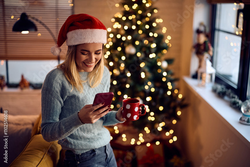 Happy woman using smart phone while having cup of tea on Christmas at home.