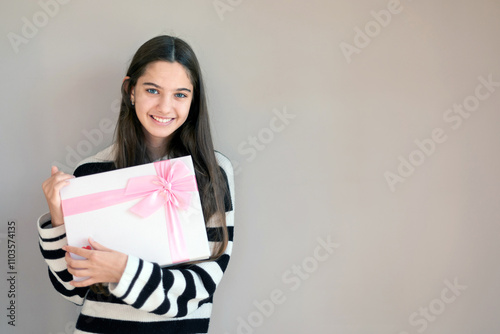 Smiling Girl Holding Gift with Pink Ribbon. Cheerful young girl with long hair holding a wrapped gift box with a pink ribbon, posing against a plain background.