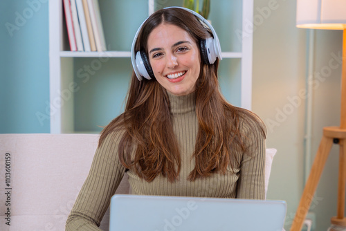 Young woman working from home with a laptop and headphones, smiling at the camera in a comfortable home office setting. Perfect for concepts of remote work, technology, and productivity.