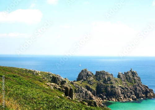 A rocky cape covered with green grass and meadow flowers against the backdrop of the endless blue ocean with a snow-white sailing ship visible in the distance, illuminated by the bright summer sun.