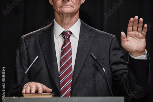 Cropped shot of senior male politician in formal attire raising hand taking oath to obey Constitution, while standing at podium on stage against of black curtains