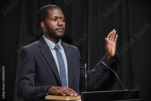 African American male politician dressed in blue suit keeping one hand on book and another raised swearing to obey law while standing at podium on stage against black curtains, copy space