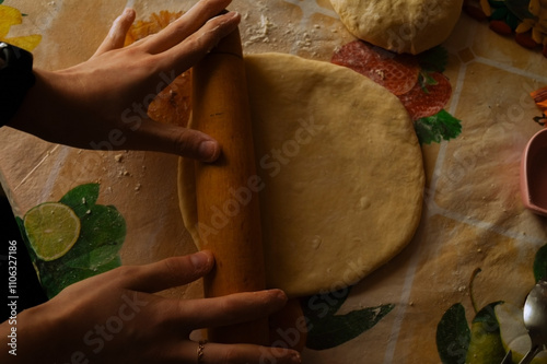 A pair of hands roll out dough with a wooden rolling pin on a vibrant, fruit-patterned tablecloth. Flour dusted on the surface adds texture to the scene, conveying a cozy baking atmosphere