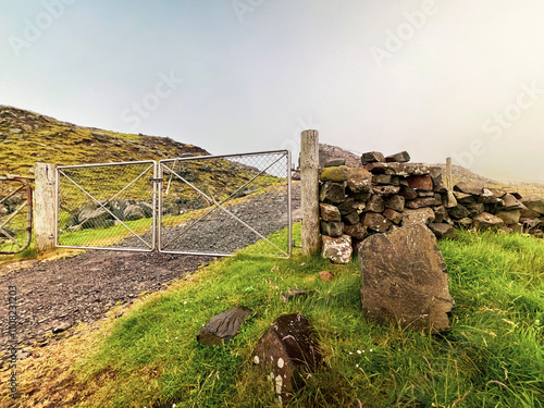 Rocky and rugged mountain trail leading uphill. A closed chain link fence run across the gravel road. A stone wall and grassy green hills surround the scene. Nólsoy, Faroe Islands.