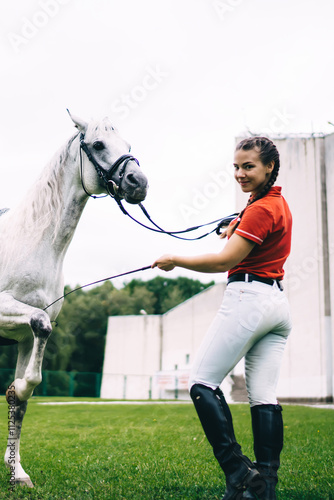 Full length portrait of Caucasian jockey girl posing during dressage competition at nature, female horseback rider looking at camera while white champion stallion animal showing stunt outdoors