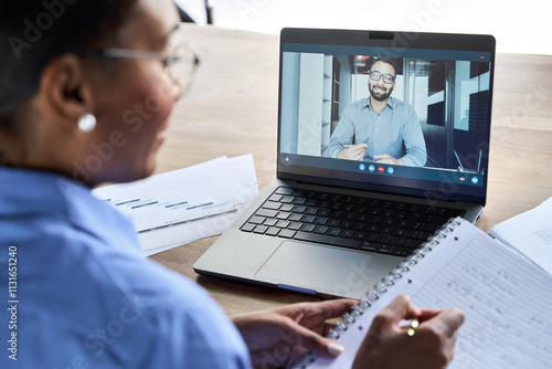 Professional African business woman manager executive working on laptop having video conference call virtual online meeting with Indian man coach, teacher, client or colleague. Over shoulder view.