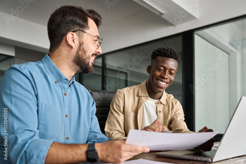 Two busy happy diverse male employees checking documents talking in office. Professional young business man manager financial advisor consulting client having conversation sitting at work meeting.