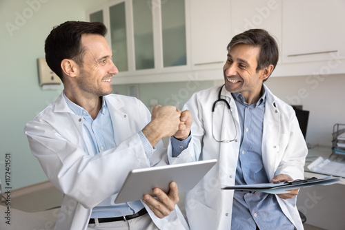 Two positive doctor colleagues making fist bump hands, enjoying successful teamwork result, analyzing medical reports together, smiling, laughing, giving friendship support gesture